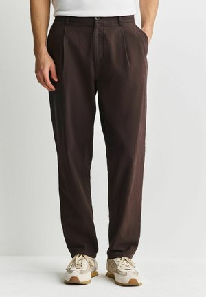 Men’s dark brown relaxed-fit trousers with button and zipper, paired with beige and white sneakers on a white background.