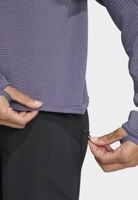 Top de manga larga texturizado en color morado con agujeros para los pulgares, combinado con leggings ajustados negros. El top cuenta con un cordón en el dobladillo para ajustarlo.