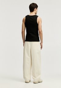 Schwarzes ärmelloses Tanktop, kombiniert mit einer lockeren, hellbeigen Hose, getragen von einer Person von hinten vor einem neutralen Hintergrund.