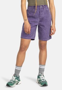 Shorts di denim viola con vita alta, tasche frontali e cuciture a contrasto; abbinate a calzini sportivi grigi e scarpe verdi.