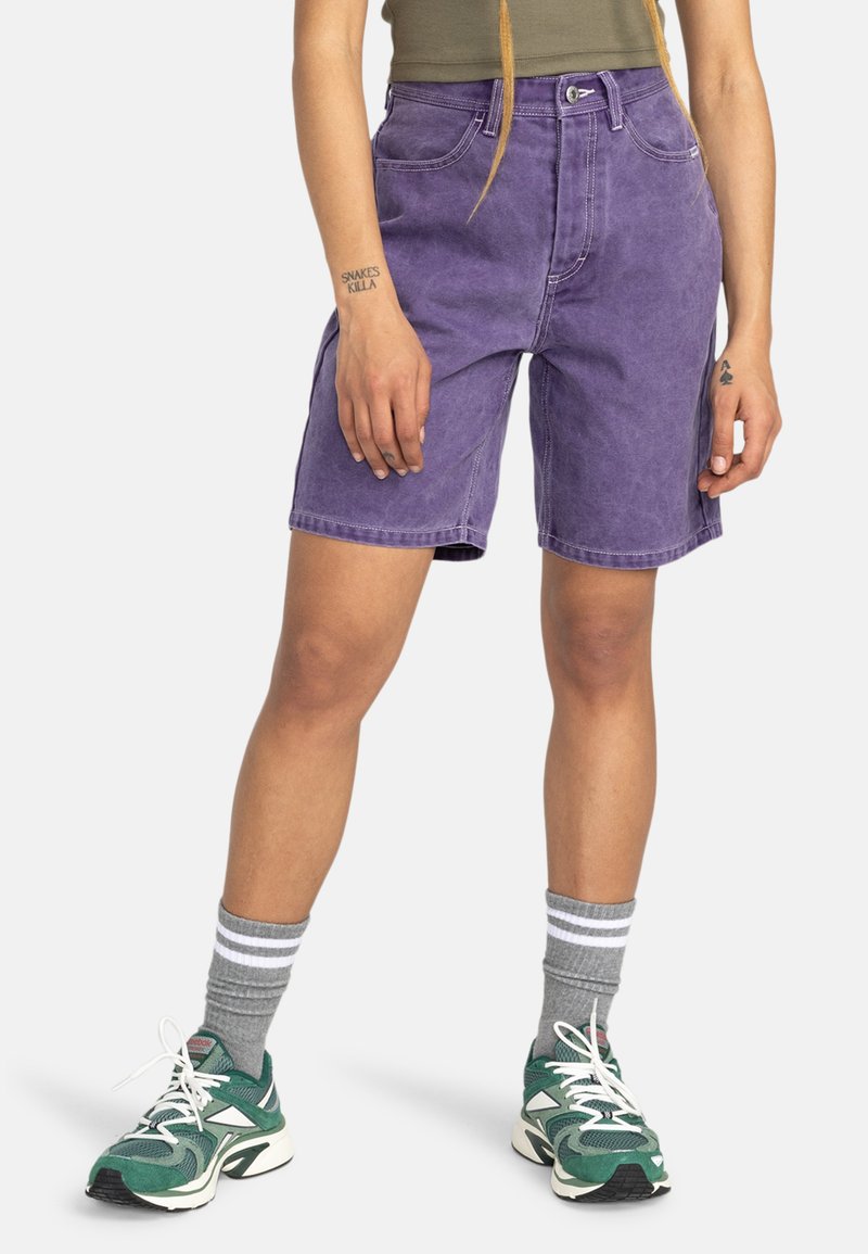 Shorts di denim viola con vita alta, tasche frontali e cuciture a contrasto; abbinate a calzini sportivi grigi e scarpe verdi.