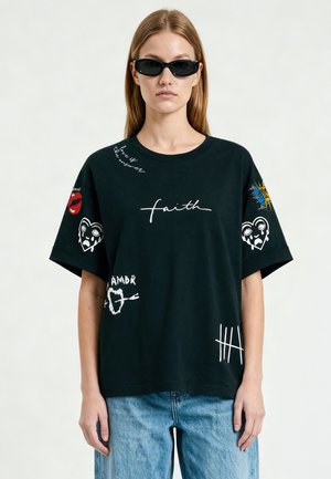 Vrouw met zwarte zonnebril, zwart oversized t-shirt met witte en kleurrijke grafische prints en een spijkerbroek tegen een effen achtergrond.