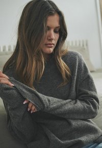 Sivý pletený sveter s voľným strihom a dlhými rukávmi, s rebrovanými manžetami a mäkkou textúrou. Ruky modelu sú čiastočne viditeľné.