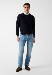 Marineblauwe trui met ronde hals over een lichtblauw overhemd met kraag; lichtblauwe spijkerbroek; bruine mocassinschoenen; model staat tegen een neutrale achtergrond.