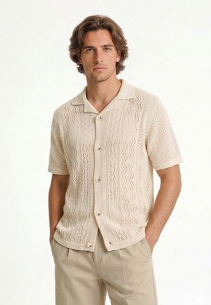 Hombre con cabello castaño de longitud media, que lleva una camisa de punto beige de manga corta con botones y pantalones beige claro con las manos en los bolsillos.