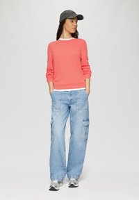 Korallenfarbener Pullover mit strukturiertem Stoff und weißem Kragen, kombiniert mit hellblauen weit geschnittenen Cargo-Jeans mit seitlichen Taschen und silbernen Laufschuhen.