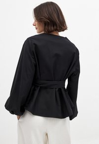 Top peplum negro con mangas largas, diseño estructurado y cintura anudada. Fabricado con tejido suave, con una textura lisa sin patrones.