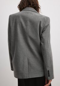 Harmaa villatakki, jossa on viistetty kaulus, pitkät hihat ja kolme nappia hihoissa. Pitkä, muotoiltu siluetti.