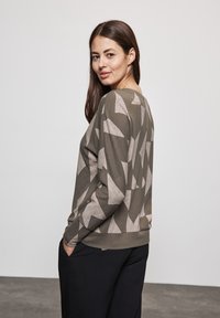 Sweatshirt à manches longues de couleur vert olive avec un motif géométrique marron clair, col rond et coupe décontractée. Pantalons noirs visibles au niveau de la taille.