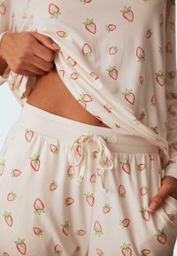 Pitkähihainen pyjaman yläosa ja housut vaaleassa kermassa, joissa on toistuvia punaisia mansikkapainanteita. Pehmeä kangas, jossa on säädettävä vyötärö.