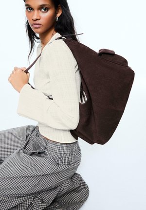 Jeune femme en haut blanc texturé et jupe à carreaux agenouillée en tenant un grand sac à dos en suède marron foncé sur une épaule.