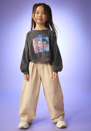Young girl standing with loose beige pants, white sneakers, and a gray sweatshirt featuring K-Pop Demon Hunters character prints.
