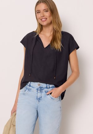Ung kvinde smiler, iført en løs sort kortærmet bluse med snore og lyseblå falmede jeans, hånd i lommen, holder beige jakke.