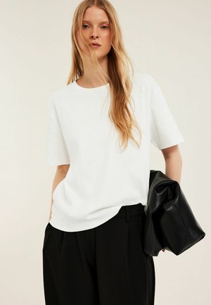 Vrouw met lang blond haar, die een wit T-shirt met korte mouwen en een zwarte broek draagt, houdt een grote zwarte leren tas onder haar arm.