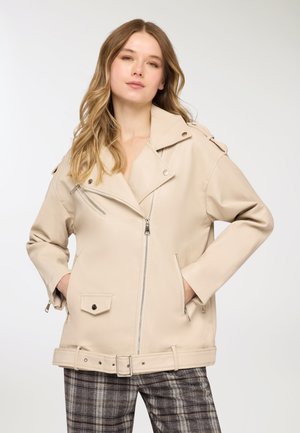 Giacca in ecopelle beige con ampi revers, tasche con zip e dettaglio con fibbia in vita. Indossata sopra pantaloni a quadri decorati.