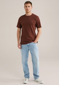 Braunes T-Shirt mit Rundhalsausschnitt und kurzen Ärmeln, kombiniert mit hellblauen Jeans. Charakteristisches Logo auf der Brust, lässige Passform und glatte Textur.