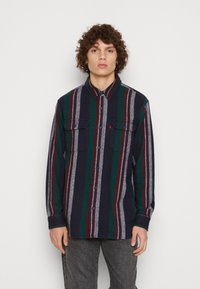 Camisa de franela a rayas en colores azul marino, verde y marrón con dos bolsillos en el pecho y cierre de botones. Textura suave y mangas largas.