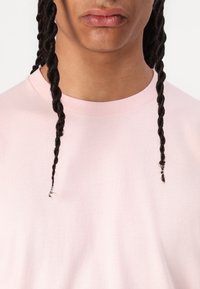 Helles rosa Baumwoll-T-Shirt mit rundem Halsausschnitt. Verfügt über eine sichtbare Textur und einen subtilen Twist im Stoff, getragen von einer Person.