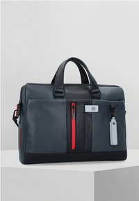 Bolso de cuero azul oscuro con acentos en cremallera roja, dos asas superiores y un parche de logo en la parte frontal. Incluye una etiqueta removible y una superficie texturizada.
