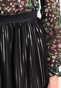 Jupe plissée noire avec une texture scintillante, dotée d'une large ceinture. Le haut présente un design floral transparent avec des fleurs colorées.