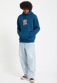Blå hættebluse i blødt stof med en lomme foran og farverigt grafisk tryk. Sammen med lyse, baggy jeans og mørke sneakers.