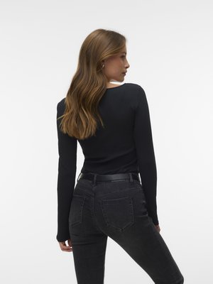 Žena s dlhými svetlohnedými vlasmi otočená mierne bokom, oblečená v priliehavom čiernom tričku s dlhým rukávom a tmavosivých džínsoch s čiernym opaskom.