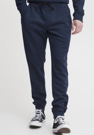 Pantalones de chándal azul marino con cinturilla elástica y cordón; textura suave; con dos bolsillos frontales y puños en los tobillos. Combinados con zapatillas negras.