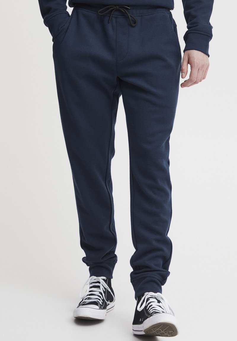 Marineblauwe sweatpants met een elastische tailleband en trekkoord; zachte textuur; voorzien van twee frontzakken en boorden aan de enkels. Gekoppeld met zwarte sneakers.