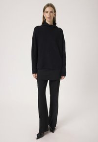 Crni, oversized pleteni pulover s visokim ovratnikom, uparen s krojenim crnim hlačama. Glatka tekstura i minimalni šavovi. Cipele na špic s mašnom.