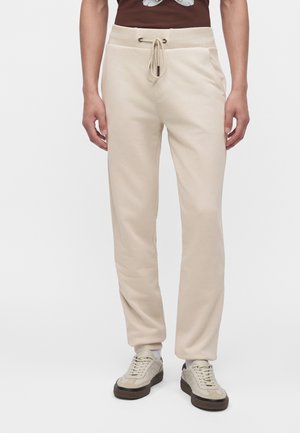 Person iført beige joggingbukser med snørre og sidelommer samt lyse beige sneakers, der står mod en ensfarvet hvid baggrund.