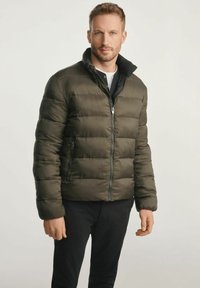 Kurtka puchowa w ciemnozielonym kolorze, z poziomym pikowaniem, wysokim kołnierzem i zapięciem na suwak z przodu. Założona na czarną koszulę i spodnie.