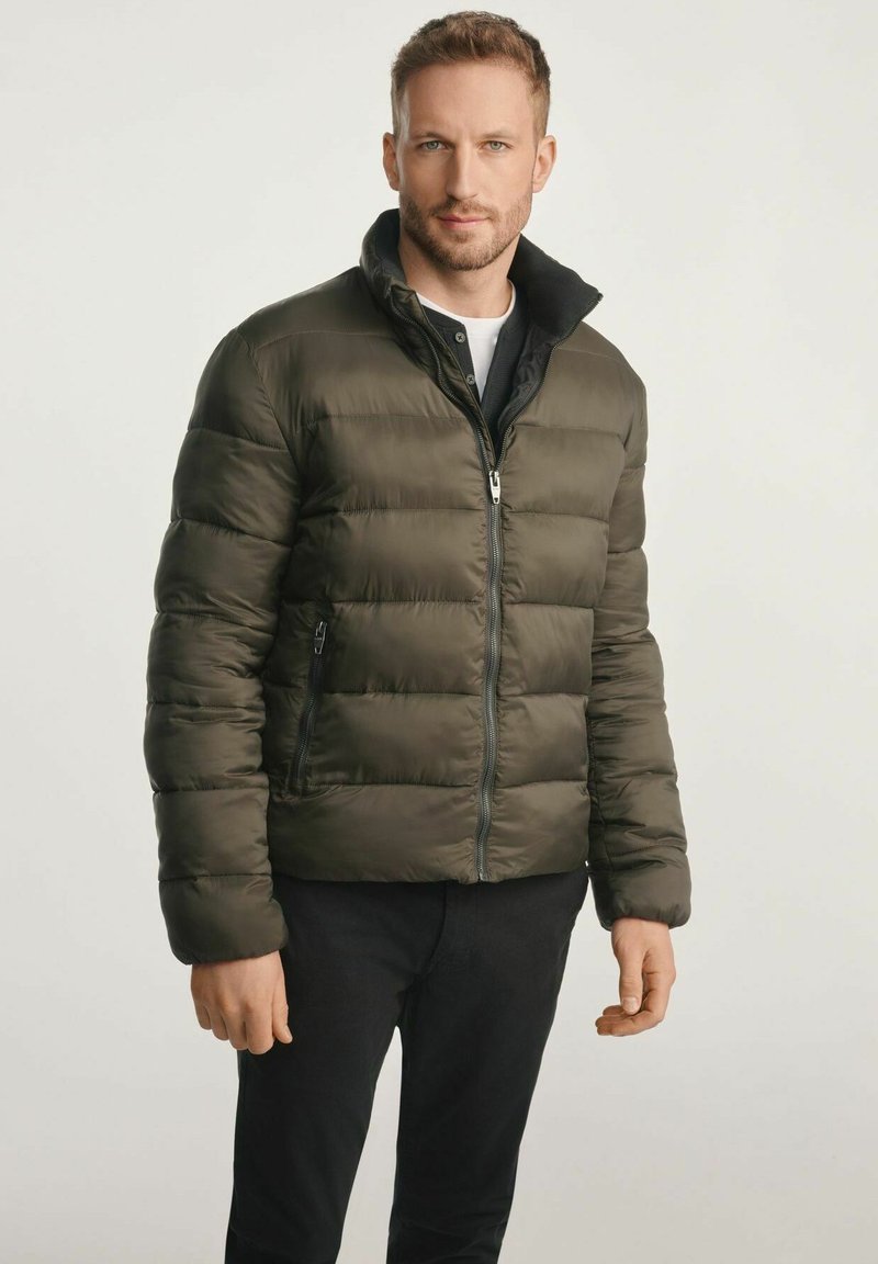 Kurtka puchowa w ciemnozielonym kolorze, z poziomym pikowaniem, wysokim kołnierzem i zapięciem na suwak z przodu. Założona na czarną koszulę i spodnie.