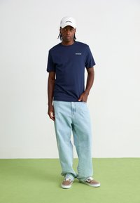 Marinblå bomulls t-shirt med kort ärm, kombinerad med ljusblå oversized jeans och bruna sneakers. Vit keps med svarta logotypdetaljer.