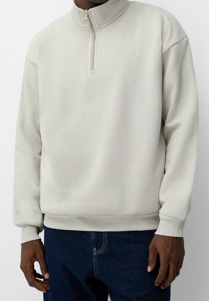 Dunkelhäutige Person trägt einen hellbeigen Half-Zip-Sweatshirt und dunkelblaue Jeans und steht vor einem schlichten weißen Hintergrund.