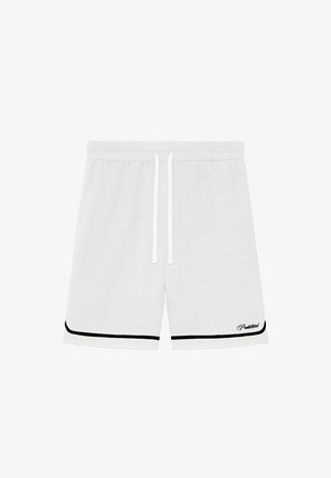 Męskie nogi w białych siateczkowych szortach z czarnym obszyciem, białe skarpetki z napisem „Prohibitions” oraz jasnobrązowe mokasyny na neutralnym tle.