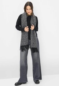 Grau-gefleckter Schal aus weichem Material mit schwarzem Fransen, getragen mit einem schwarzen Pullover und kombiniert mit weiten Denim-Hosen.