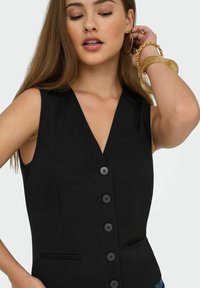 Frau in ärmellosem schwarzen, geknöpftem Weste mit tiefem V-Ausschnitt, großen Creolen und klobigen Armbändern, die ihr Haar berührt.