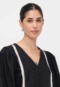 Black blouse with voluminous sleeves, lace trim down the front, and button closure. Model wears silver star-shaped earrings.