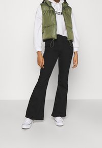 Chaleco acolchado en verde oliva sobre una sudadera blanca con capucha, combinado con jeans de campana negros y zapatillas blancas, con cierre de cremallera y dobladillo ajustable.