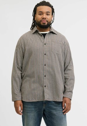 Mann med dreadlocks iført brun og hvit rutete skjorte og blå jeans, stående mot en ensfarget hvit bakgrunn.