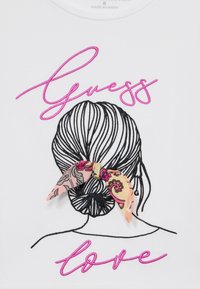 Baltas audinys su juodu siuvinėtu plaukų dizainu, kaip ir rožinėmis užrašais "Guess" ir "love"; papuoštas modeliuotu kaspinu plaukų linijoje.