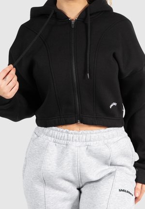 Vrouw draagt een zwart, kort zip-up hoodie en lichtgrijze sweatpants met elastische taille, houdt een trekkoord van de hoodie vast.