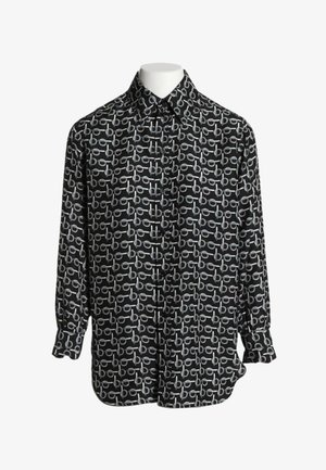 Zwarte blouse met lange mouwen en witte repeterende patroon van gekoppelde cirkels en horizontale balken, getoond op een hoofdloze paspop.