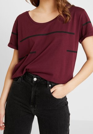 Femme portant un t-shirt bordeaux à manches courtes avec des rayures horizontales noires et un jean noir taille haute, main dans la poche.