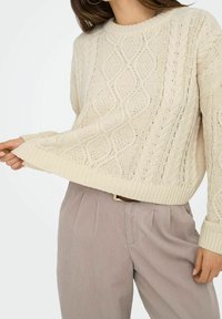 Cremefarbener Strickpullover mit strukturiertem Zopfmuster, gerippten Bündchen und Saum. Kombiniert mit hellgrauen, plissierten Hosen.