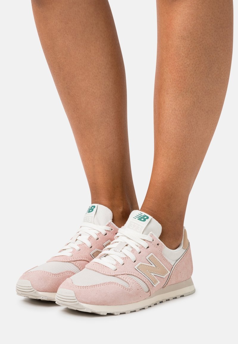 Jambes portant des baskets New Balance roses et blanches avec des accents beige et un logo vert, debout sur une surface blanche.