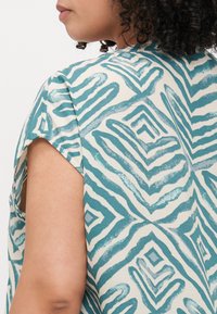 Loose-fitting short-sleeve top in cream with teal geometric patterns; features a V-shaped neckline and soft, textured fabric.