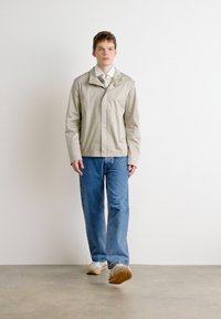 Lys grå jakke med krage, kombinert med blå jeans og hvite joggesko. Modellen står foran en nøytral bakgrunn. Jevn tekstur, enkelt design.