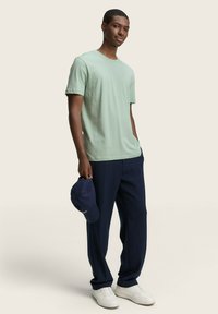 T-shirt in cotone verde chiaro abbinato a pantaloni blu navy, sneakers bianche e un cappellino blu navy tenuto nella mano destra. Design semplice e texture morbida.