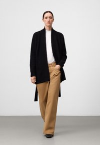 Svart stickad cardigans med öppen front, parat med raka beige byxor. Enkel vit t-shirt under, mot en neutral bakgrund.